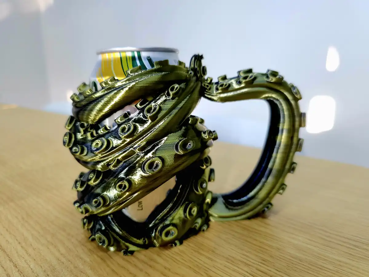 A 3D-printed, bronze-colored octopus tentacle holder grips a beverage can on a wooden surface. The tentacles have detailed…