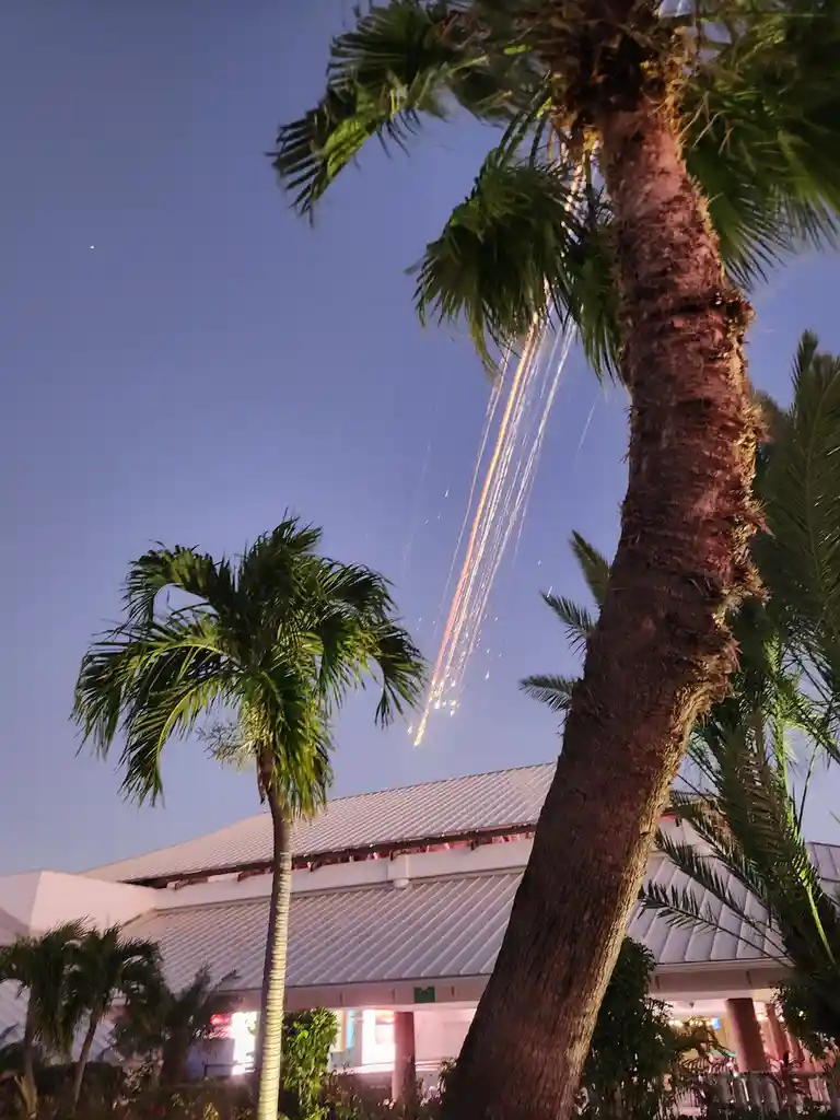 A low-angle nighttime photo shows palm trees in front of a building, with bright streaks of orange and white fireworks…