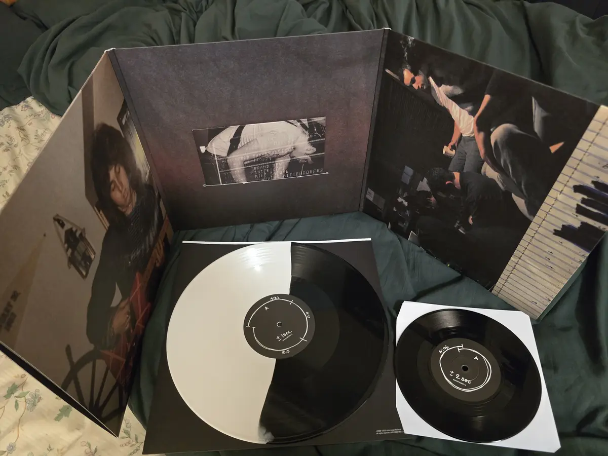 An open vinyl record album lies on a green bedspread, revealing a split record with one half white and one half black. The…