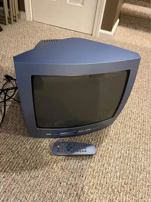 A vintage blue CRT television sits on a beige carpet, with a matching remote control placed in front of it. The TV screen is…