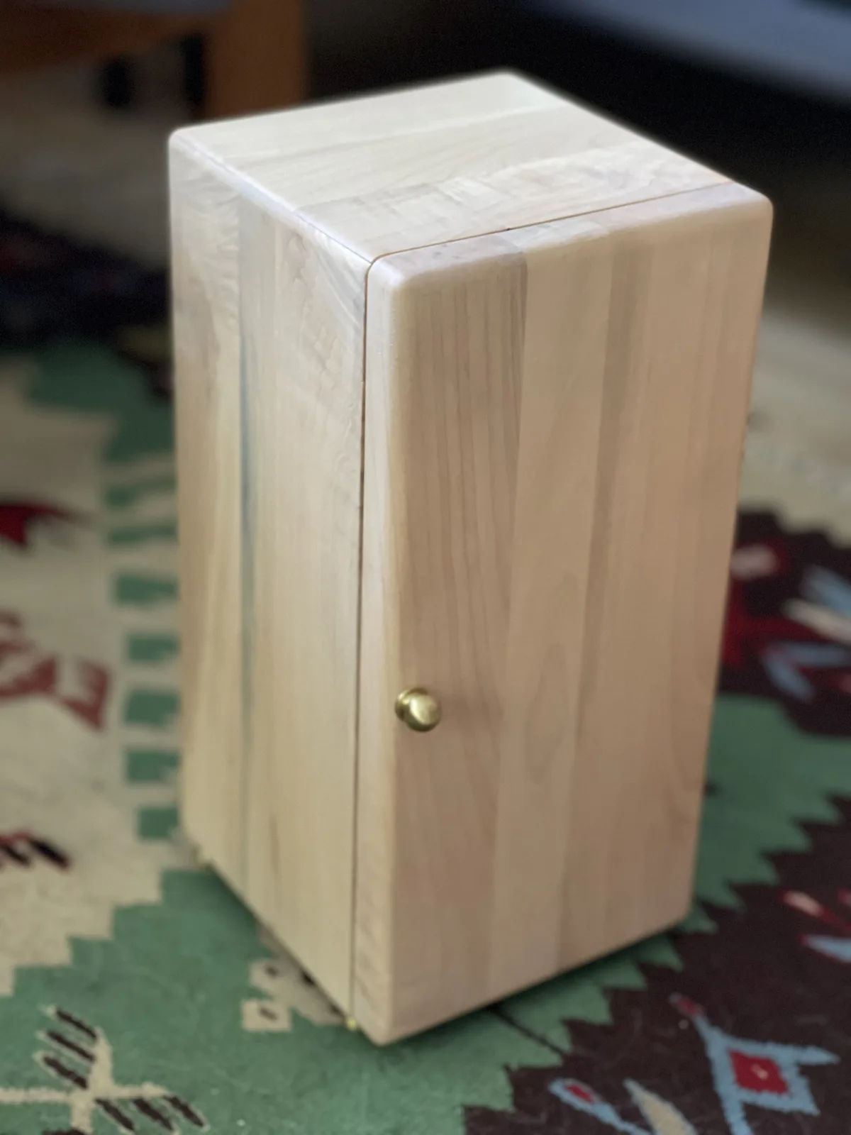 A light-colored wooden box with a small brass knob on its side sits on a patterned rug. The box appears handmade and has a…