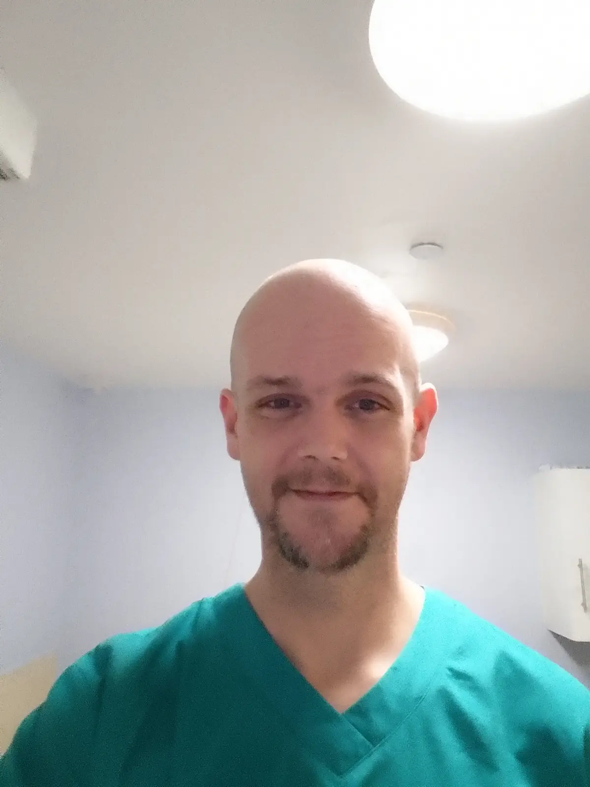 A bald man with a goatee wearing green scrubs stands in a brightly lit room with a large circular ceiling light above him…