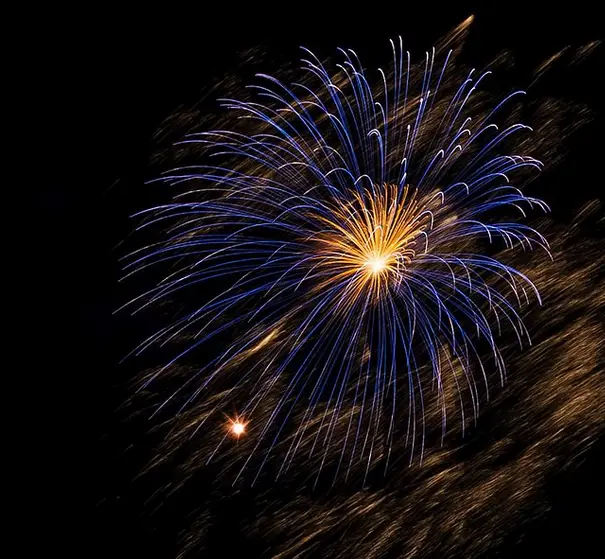 A long-exposure photograph captures a vibrant blue and gold fireworks explosion against a black night sky, with streaks of…