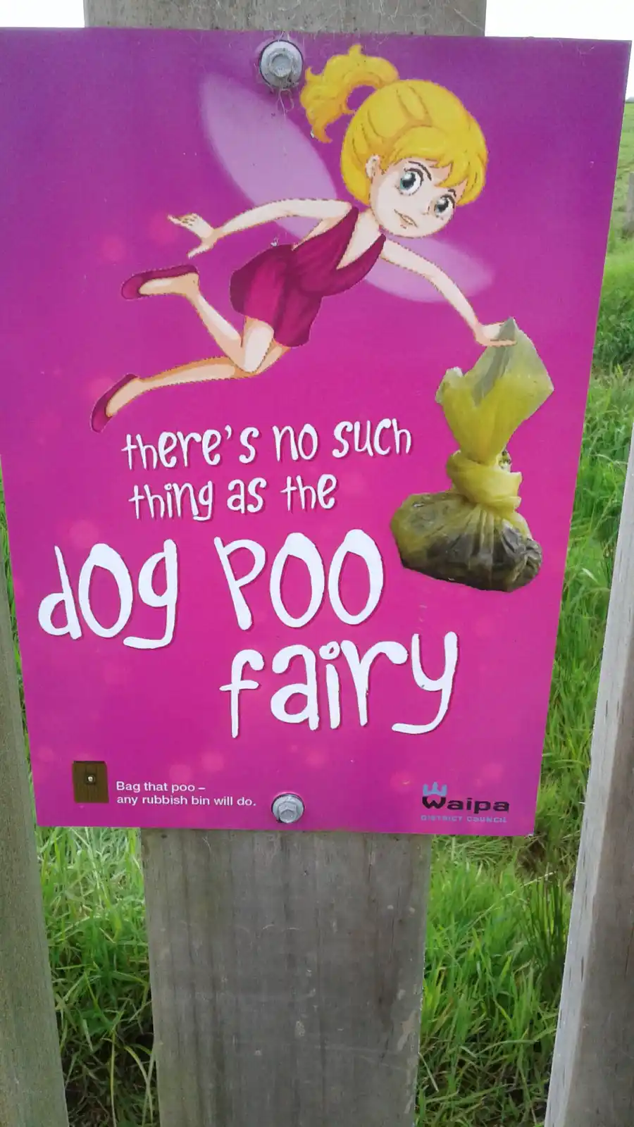 A pink public sign mounted on a wooden post features a cartoon fairy with blonde hair and purple wings, holding a bag of dog…