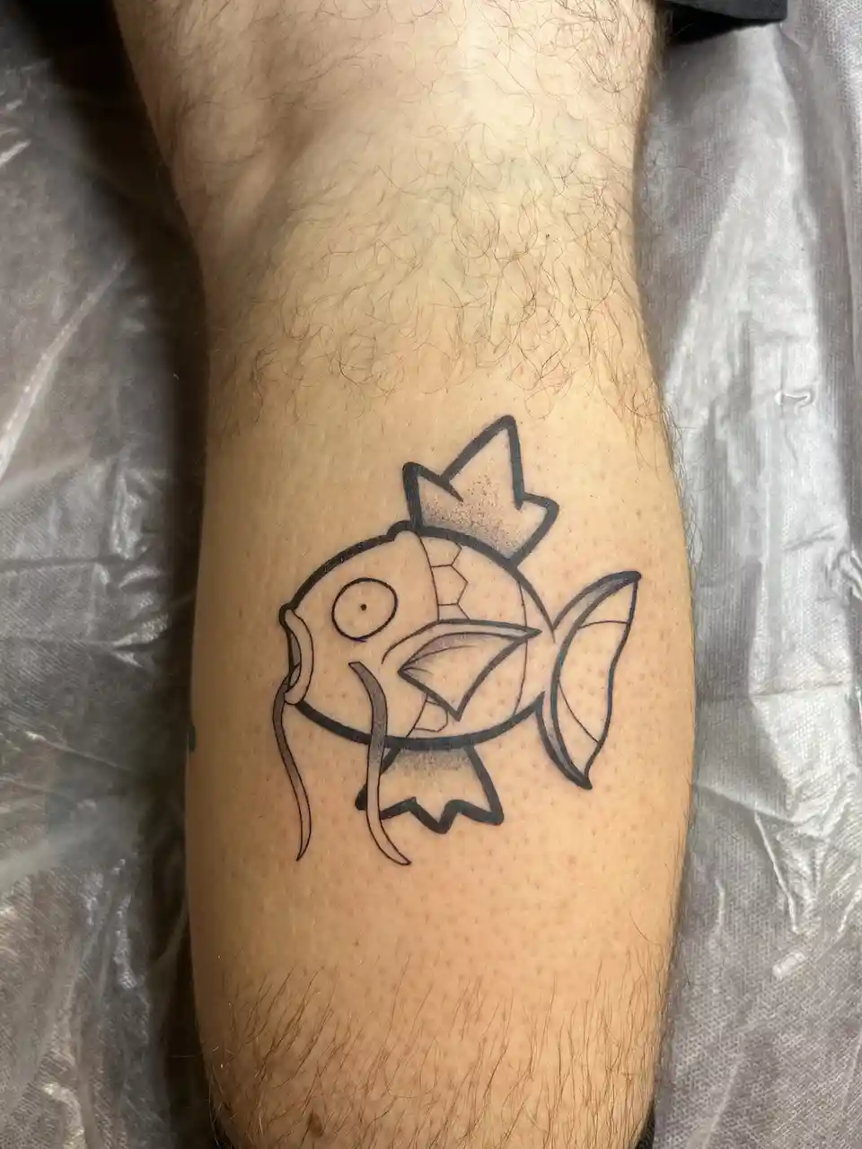 A close-up photo shows a black-and-white tattoo of the Pokémon Magikarp on a person's hairy forearm, with the text 'i got a…