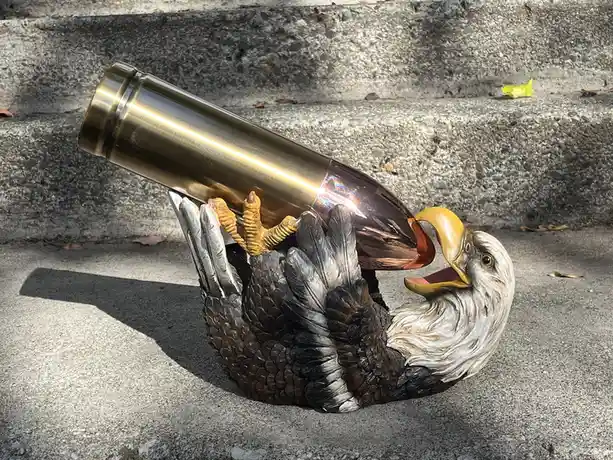 A plastic bald eagle statue lies on its back on a concrete surface, gripping a large brass-colored bullet with its talons…