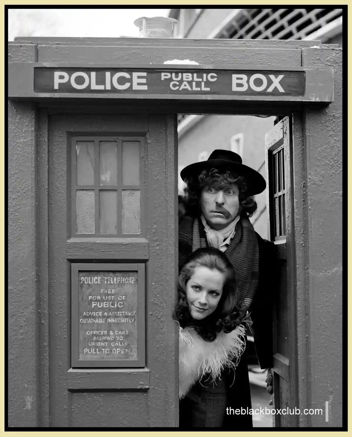 A black-and-white photo shows two people peeking out from a vintage British police public call box. The man behind has a…