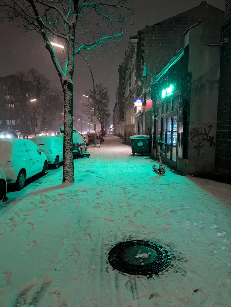 A snowy urban street at night, illuminated by a strong green glow from a storefront, with snow-covered cars parked along the…