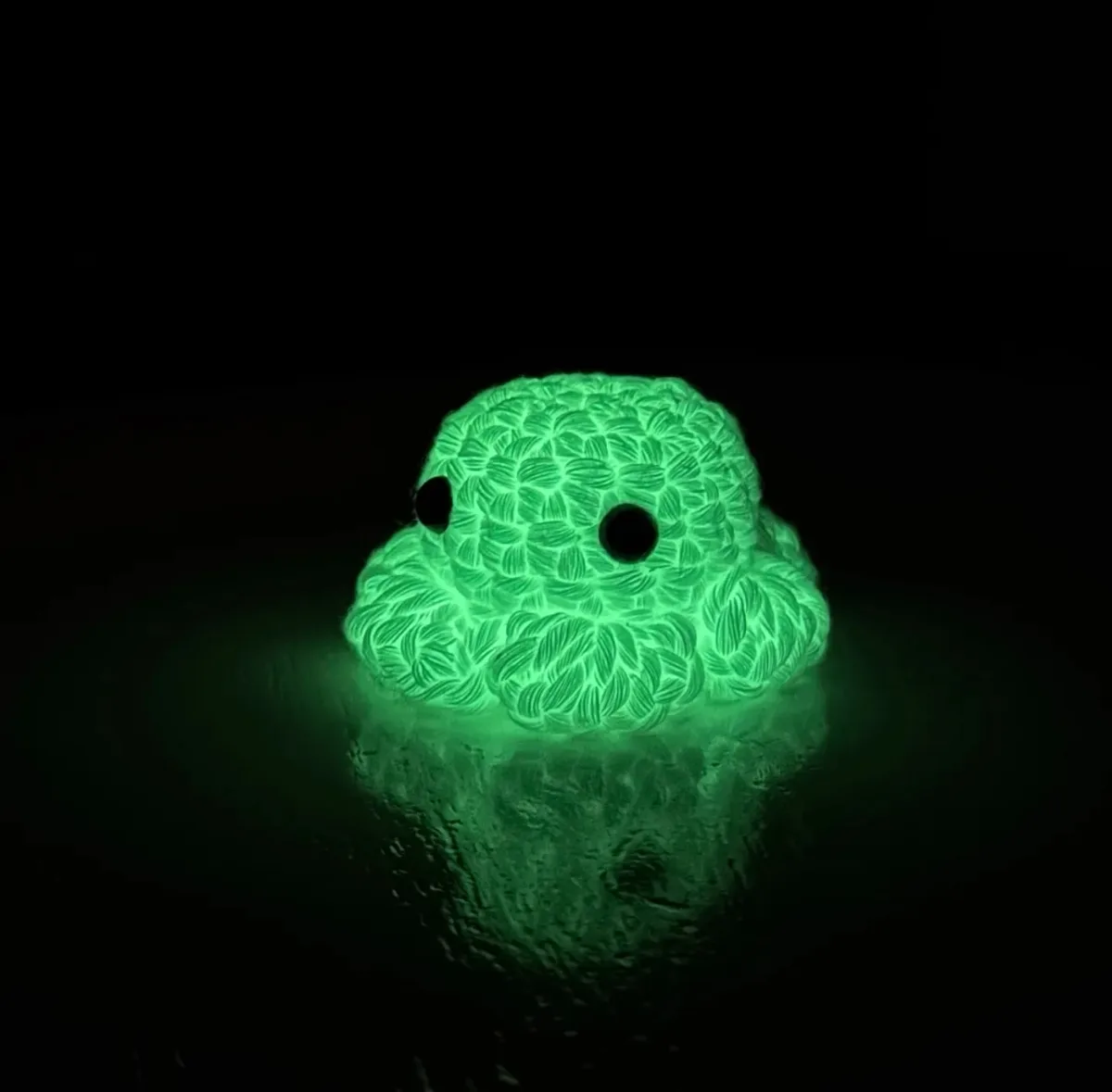 A small, glowing green knitted octopus toy with large black eyes sits in a dark, reflective surface, casting a green glow…