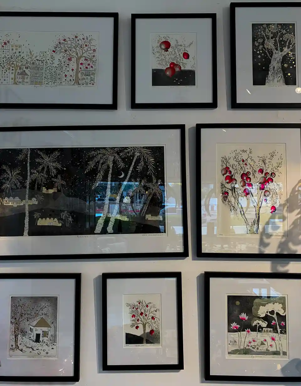 A wall displays nine framed artworks, mostly featuring stylized trees with red fruits like pomegranates or flowers, set…