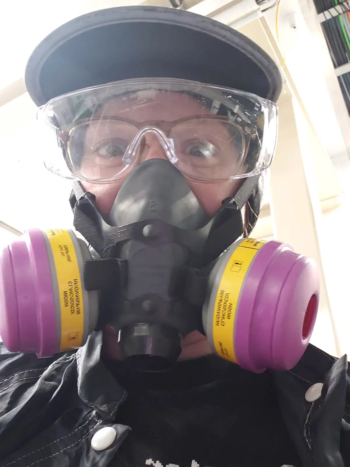 A person wearing a black baseball cap, clear safety goggles, and a full-face respirator mask with purple filter cartridges…