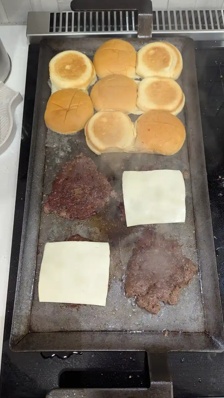 A close-up photo shows a griddle with nine toasted burger buns arranged in a row at the top, and three burger patties with…