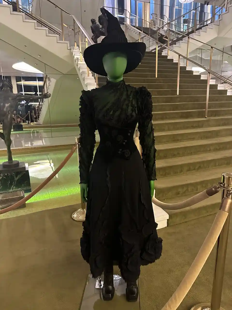 A mannequin dressed in a detailed black witch costume with a tall pointed hat and green skin stands on display in a…