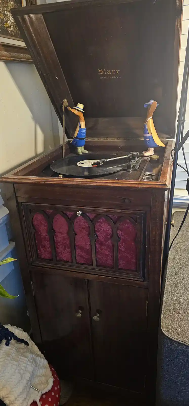 A vintage wooden record player with its lid open, featuring two small wooden duck figurines dressed in overalls and hats…