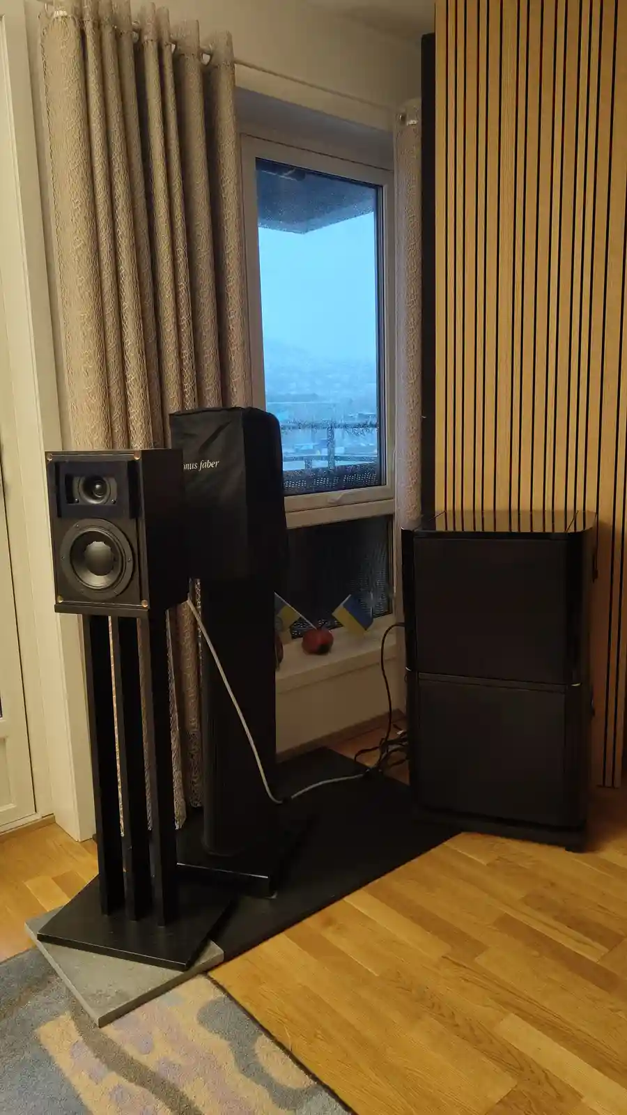 A room with two large black speakers, one on a stand and one on the floor, positioned near a window with curtains. The…