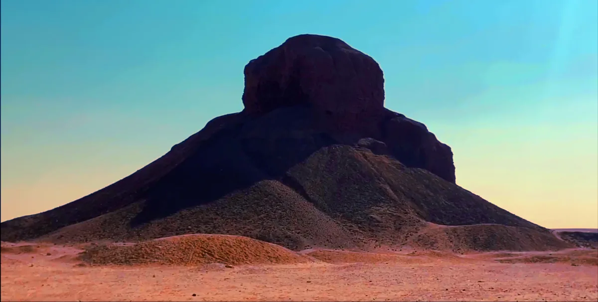 A desert landscape featuring a large, dark, pyramid-shaped rock formation under a gradient sky transitioning from blue to…