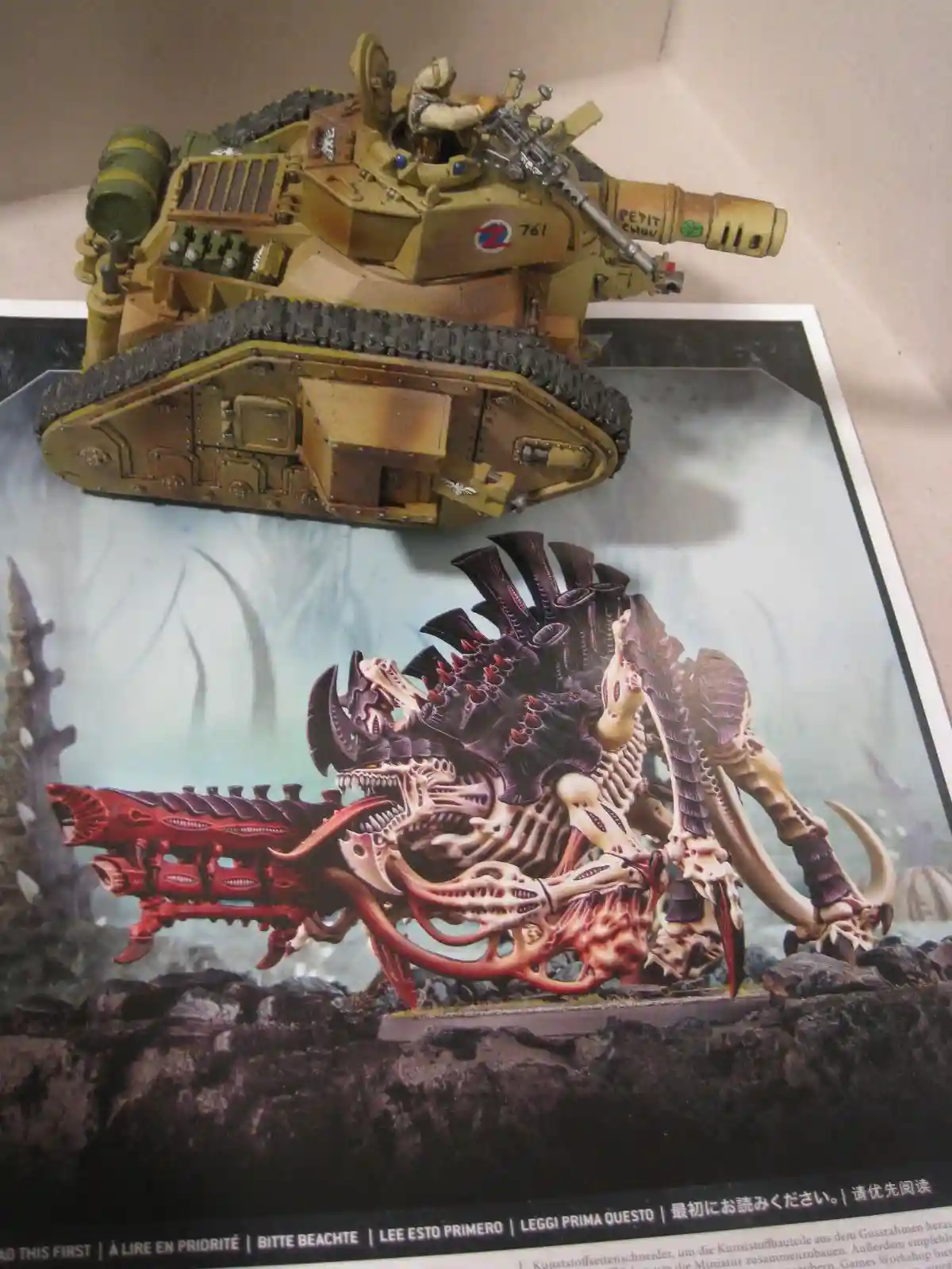 A painted miniature of a tan, heavily armed tank sits atop a printed image of a monstrous, biomechanical alien creature with…