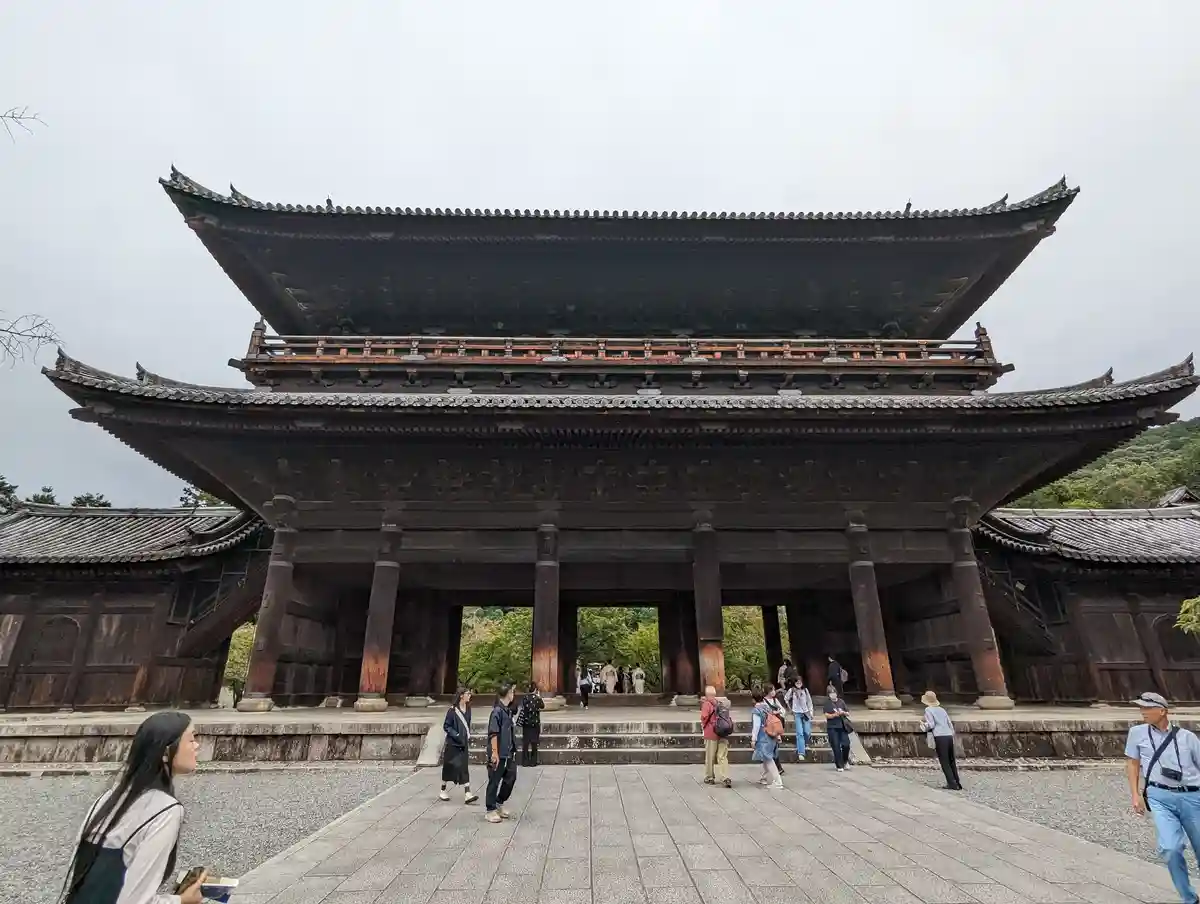 A large, traditional Japanese wooden gate with multiple tiers and curved roofs stands under an overcast sky, with several…