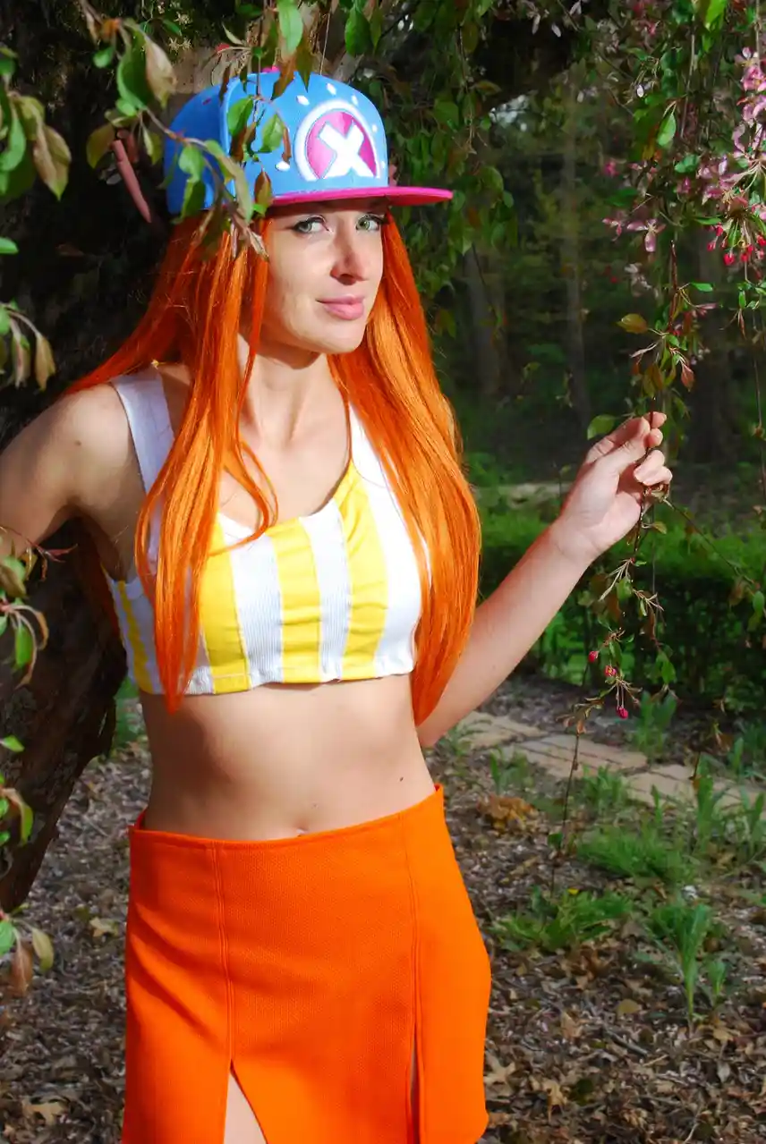 A person with long orange hair and green eyes is cosplaying as the character Ash Ketchum from Pokémon, wearing a blue and…