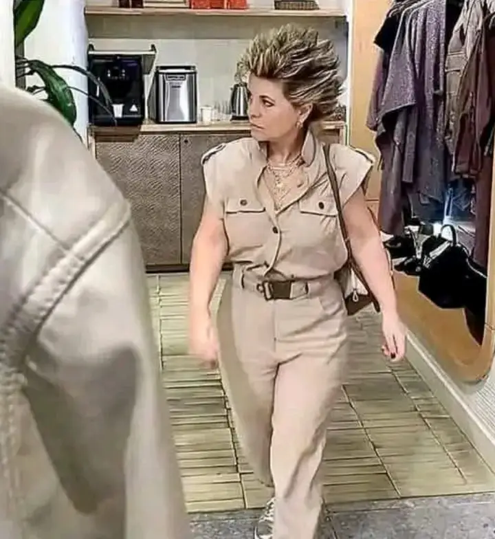 A woman with a spiky, messy hairstyle walks forward in a beige jumpsuit, looking annoyed or unimpressed. She carries a purse…