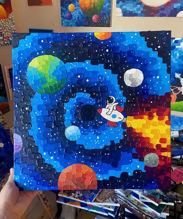 A hand holds up a textured, thickly painted canvas depicting a colorful space scene with planets, stars, and an astronaut in…