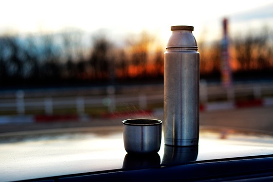 A silver thermos and a small metal cup sit on the hood of a car during sunset, with a blurred background of trees and a…