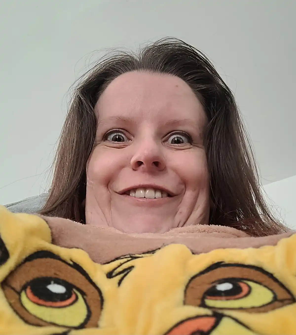 A person with brown hair and wide, surprised eyes smiles while peeking over a yellow, plush blanket featuring large cartoon…