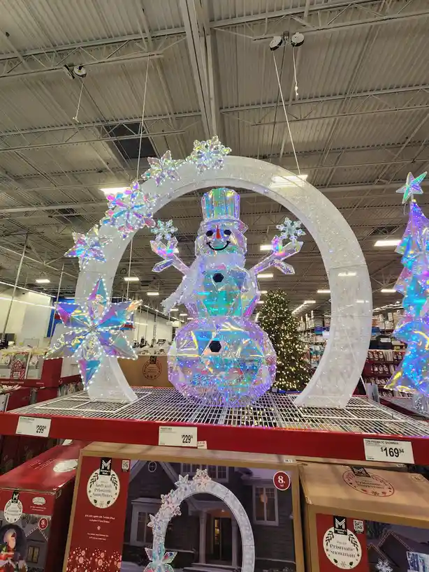 A large, illuminated, prismatic snowman decoration is displayed in a retail store aisle, suspended under a glittering arch…