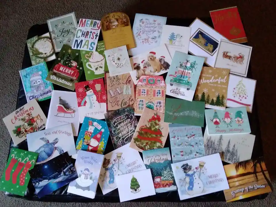 A large pile of diverse Christmas greeting cards is spread across a dark surface, featuring various holiday-themed…