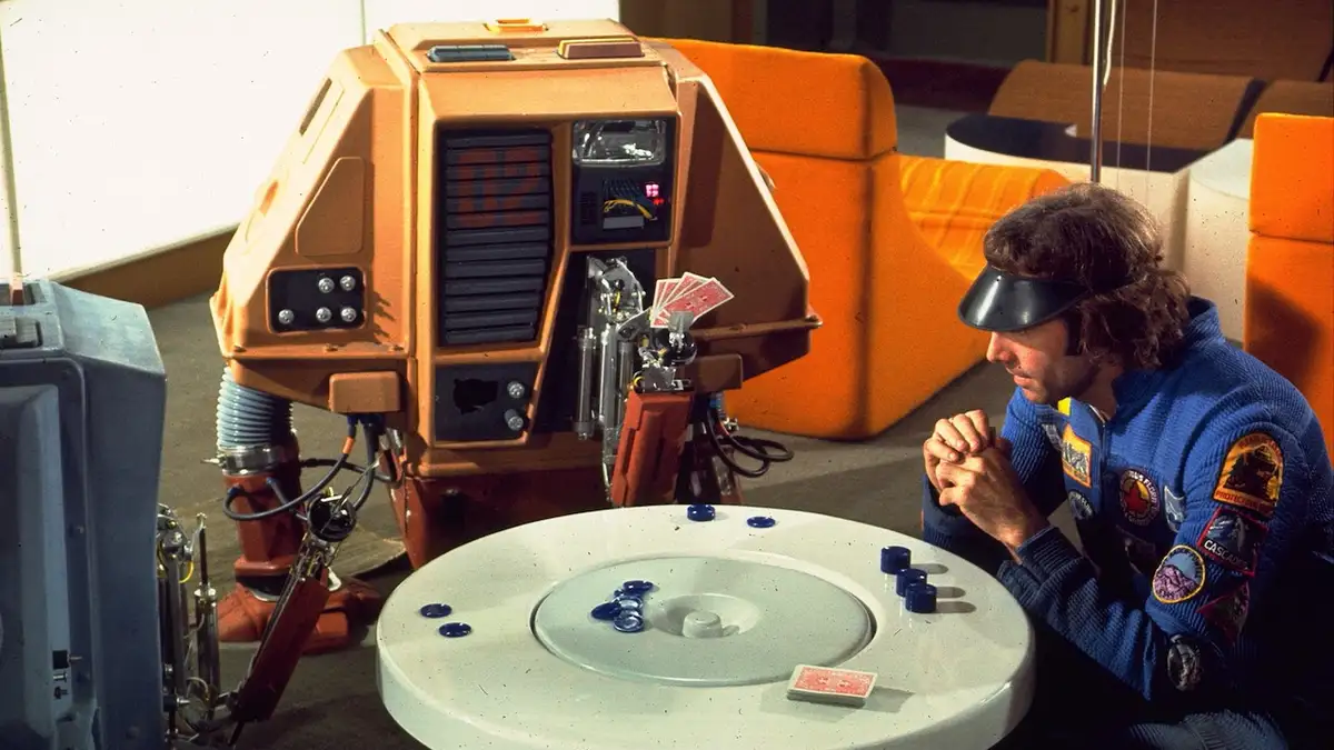 A man in a blue astronaut-style jumpsuit with patches and a black visor sits at a white circular table, clasping his hands…