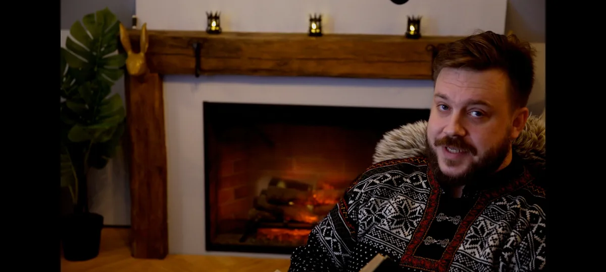A man with a beard and mustache wearing a patterned sweater with a fur-lined hood sits in front of a fireplace. He looks…
