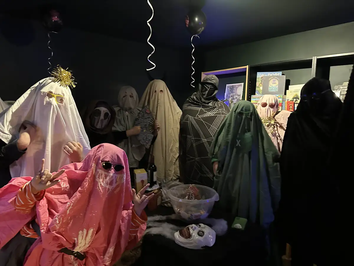A group of people dressed in various ghost costumes, including a pink one in the foreground making hand gestures, stand in a…