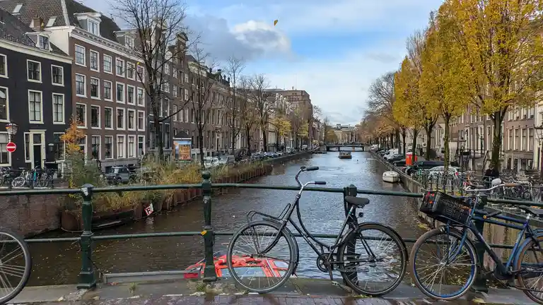 A scenic autumn view of a canal in Amsterdam with historic buildings, bare and yellow trees, and bicycles parked on a bridge…