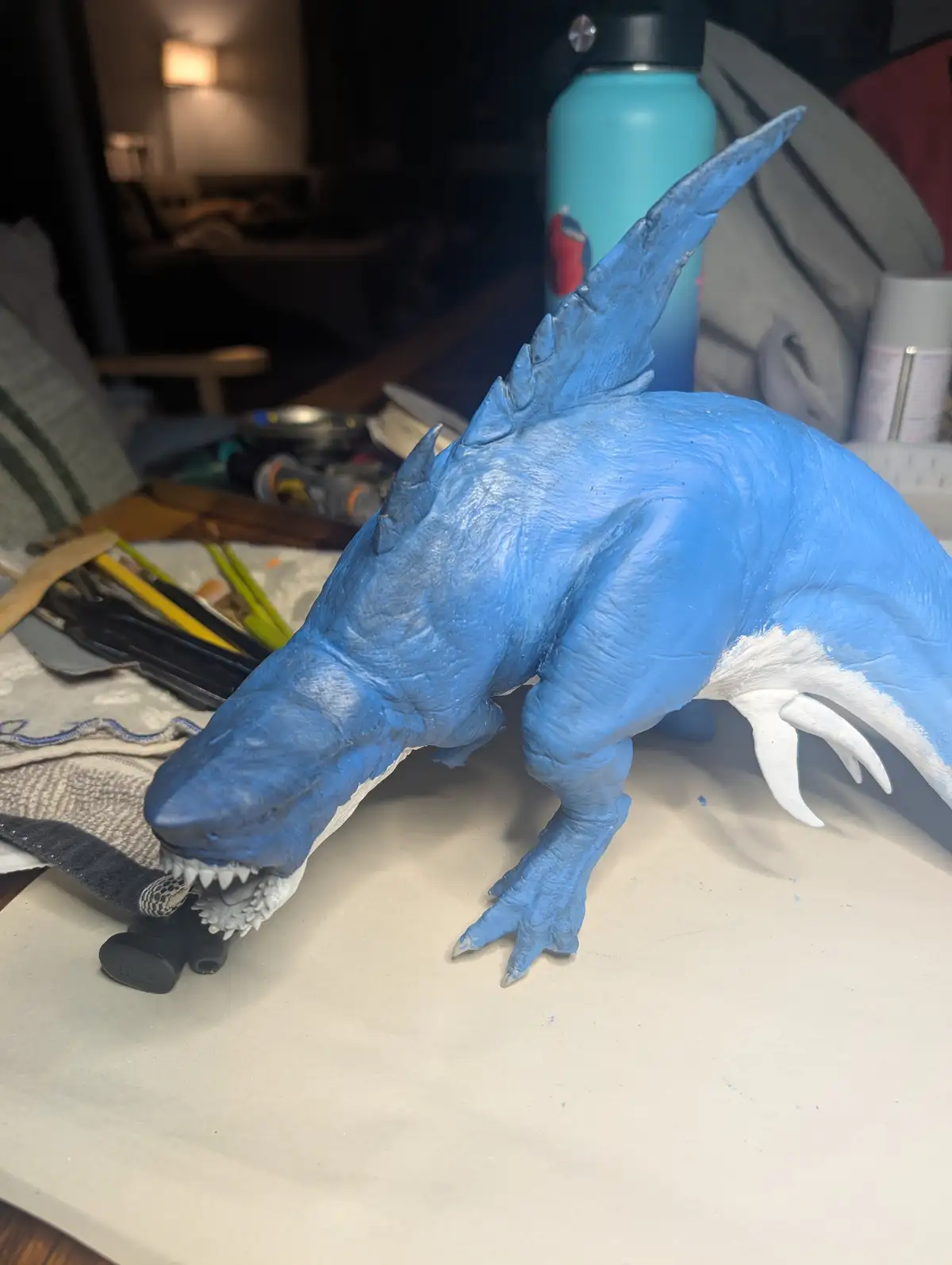 A blue-painted dragon sculpture with white accents on its underbelly and claws sits on a workbench, surrounded by art…