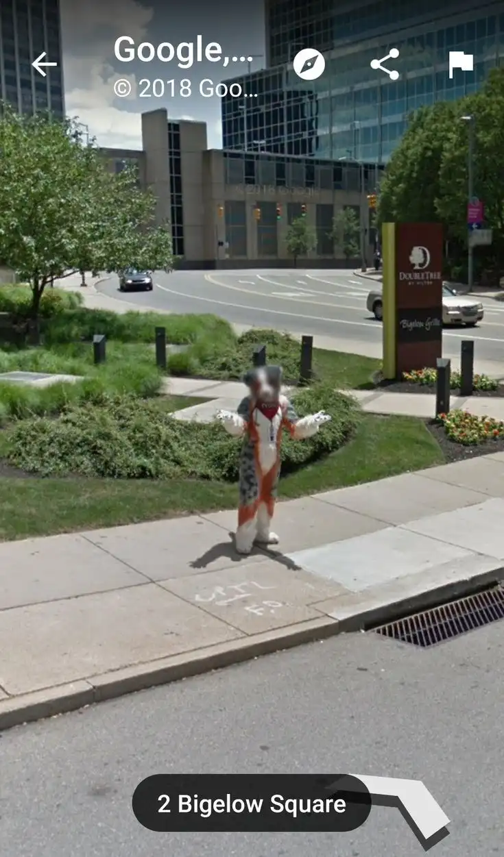 A person in a full-body cat mascot costume stands on a sidewalk in front of a DoubleTree hotel, with a Google Street View…
