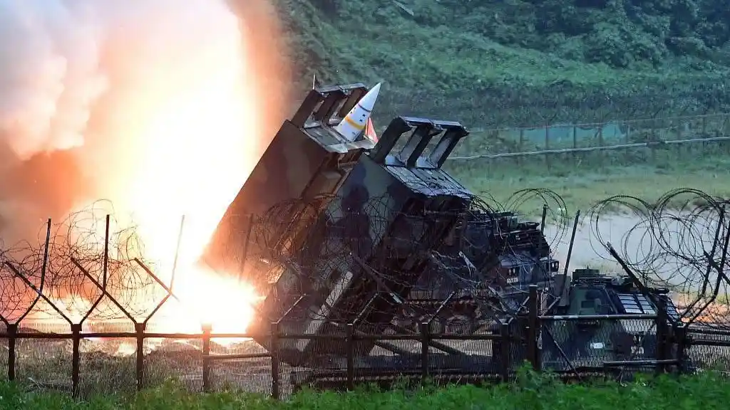 A military rocket launcher fires a missile, creating a bright explosion and smoke, with barbed wire fencing in the…