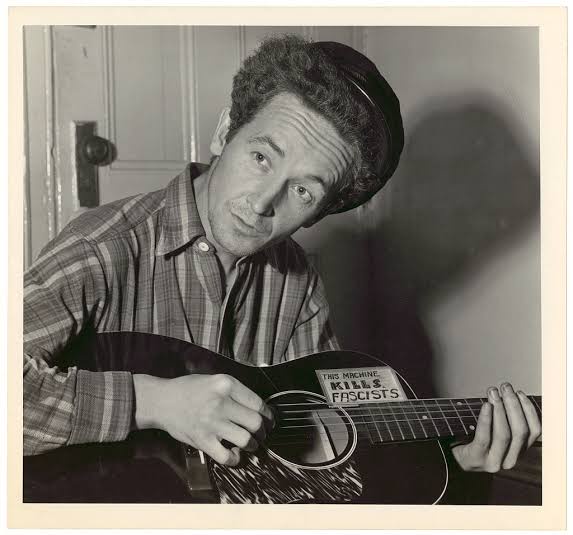 A black-and-white photo of a man with curly hair wearing a cap and plaid shirt, holding an acoustic guitar. A sticker on the…