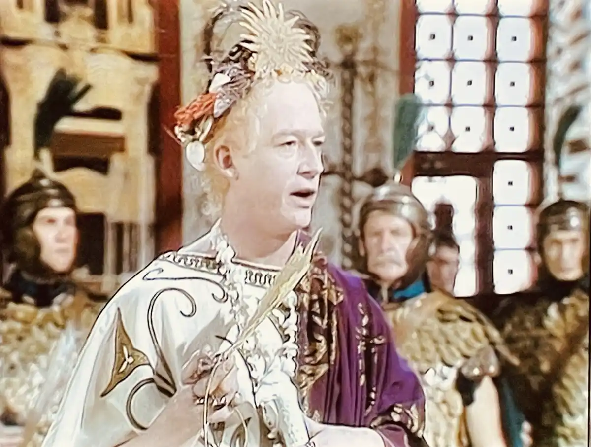 A man in elaborate historical royal attire, including a jeweled crown and ornate robes, stands in a regal setting with…