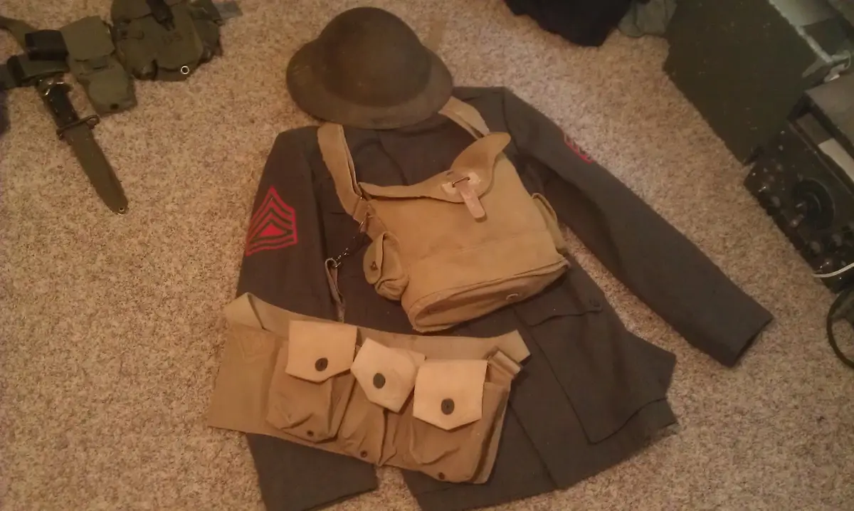A collection of World War I-era U.S. military gear, including a dark uniform jacket with red sergeant stripes, a helmet, a…