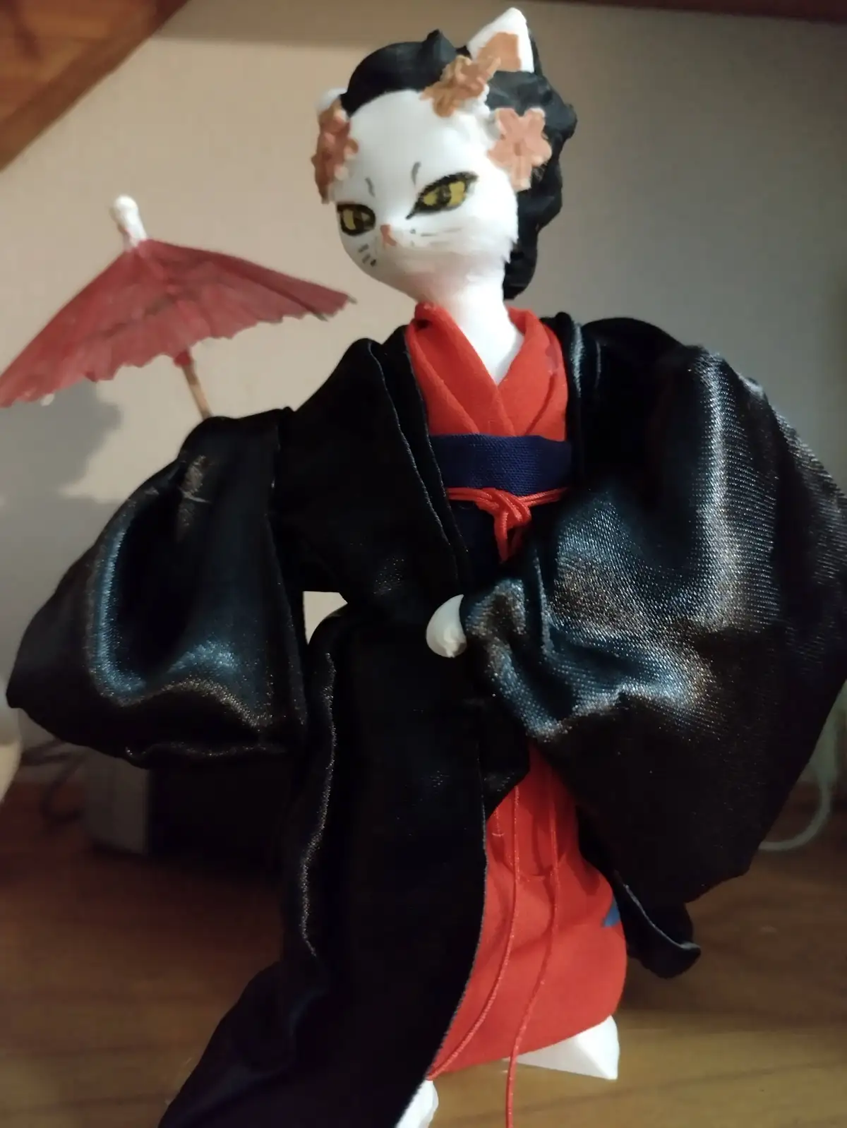 A handmade cat figurine dressed in traditional Japanese geisha attire, including a black outer robe over a red undergarment…