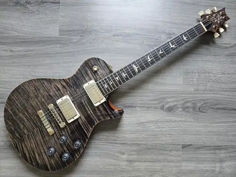 An electric guitar with a dark wood grain finish and bird-shaped inlays on the fretboard lies diagonally on a gray wood…