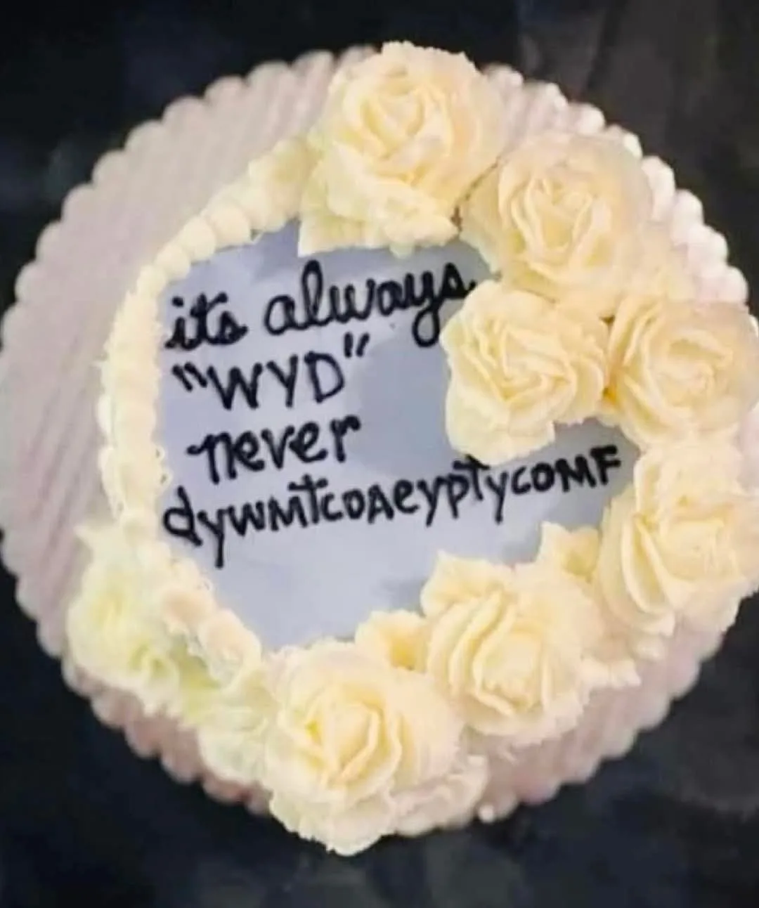A round cake with white frosting roses around the edge and a handwritten message in black icing on the center. The text…