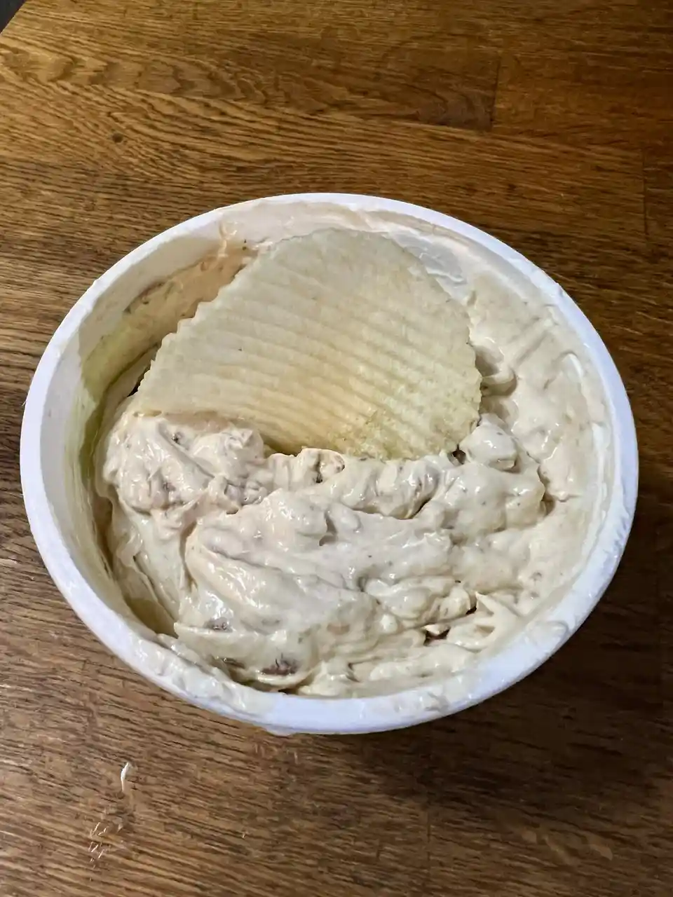 A top-down photo shows a single ridged potato chip resting in a white plastic tub filled with creamy, chunky dip, placed on…