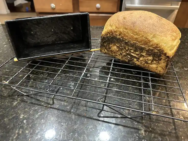 A loaf of bread with a heavily burnt bottom sits on a wire cooling rack next to an empty black loaf pan, both resting on a…
