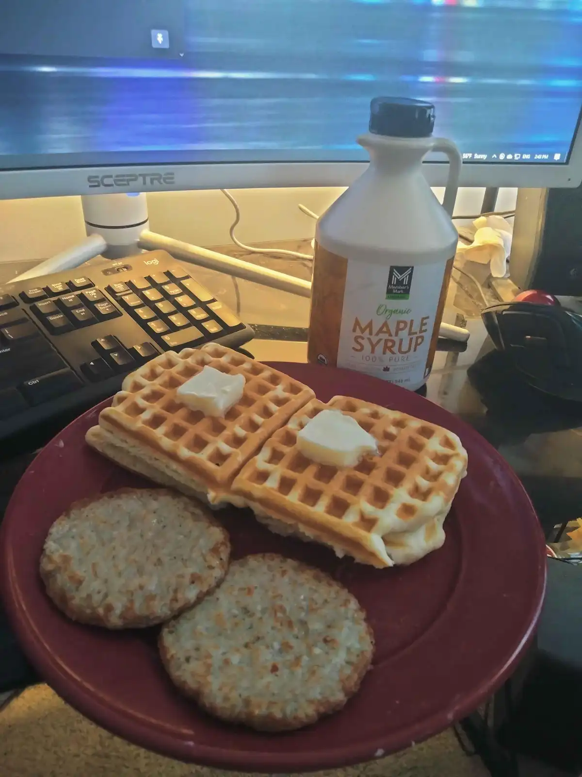 A red plate holds two buttered waffles and two sausage patties on a cluttered desk, with a computer monitor, keyboard, and a…