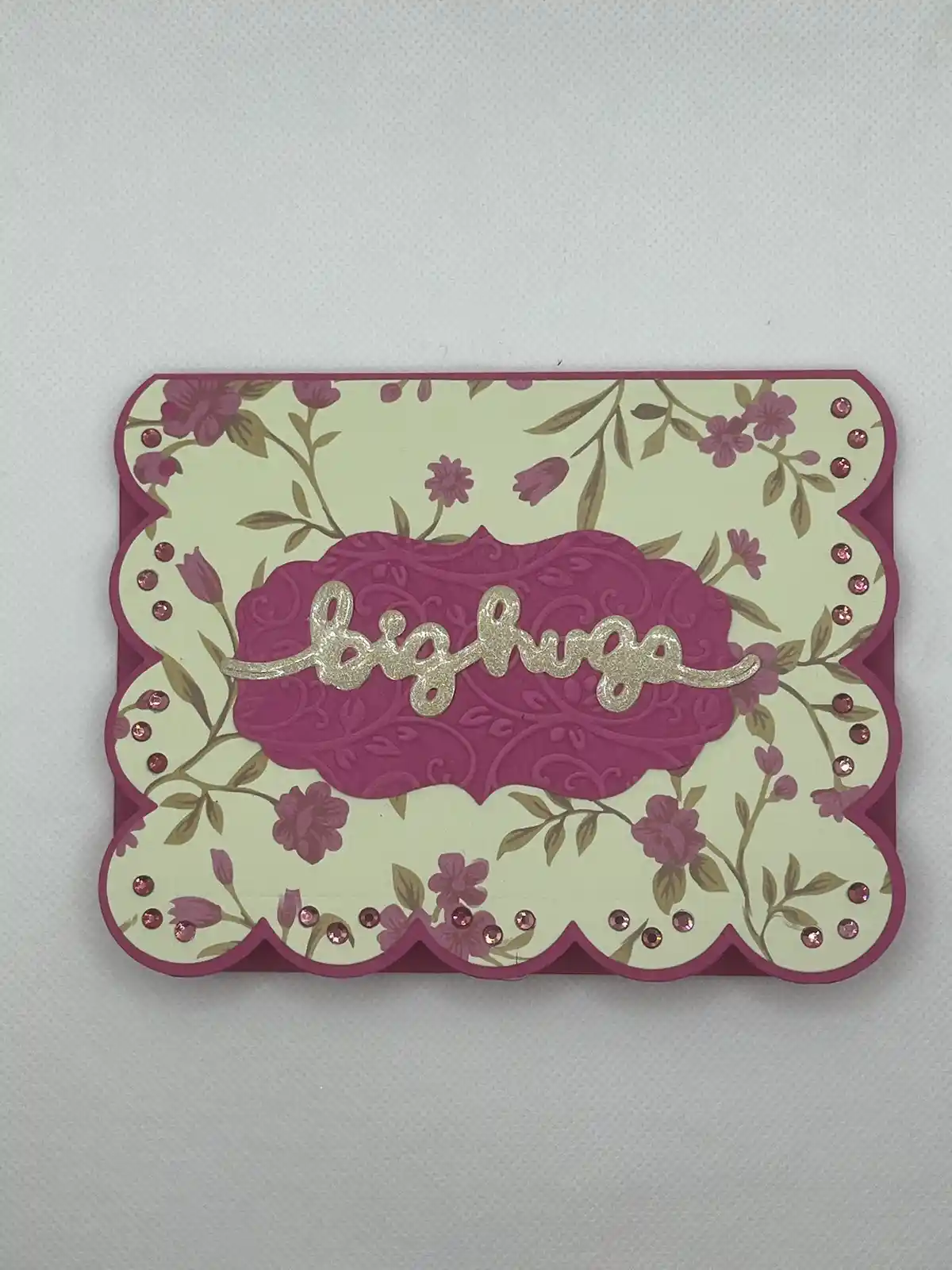 A handmade greeting card with a scalloped magenta border and floral patterned background, adorned with pink rhinestones. The…