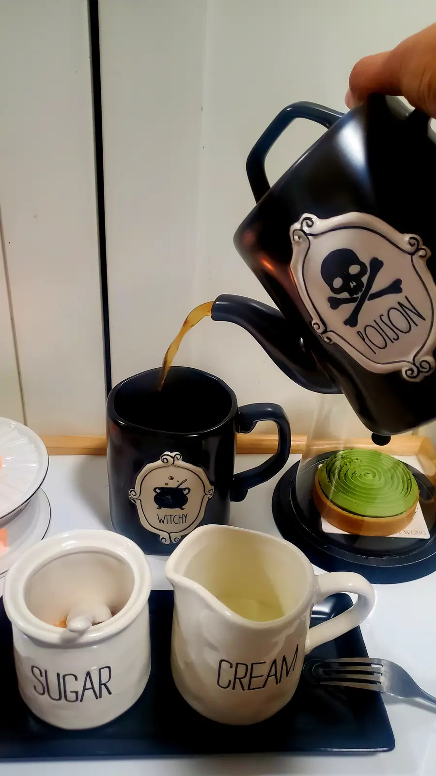A hand pours coffee from a black teapot labeled 'POISON' with a skull and crossbones into a black mug labeled 'WITCHY'…