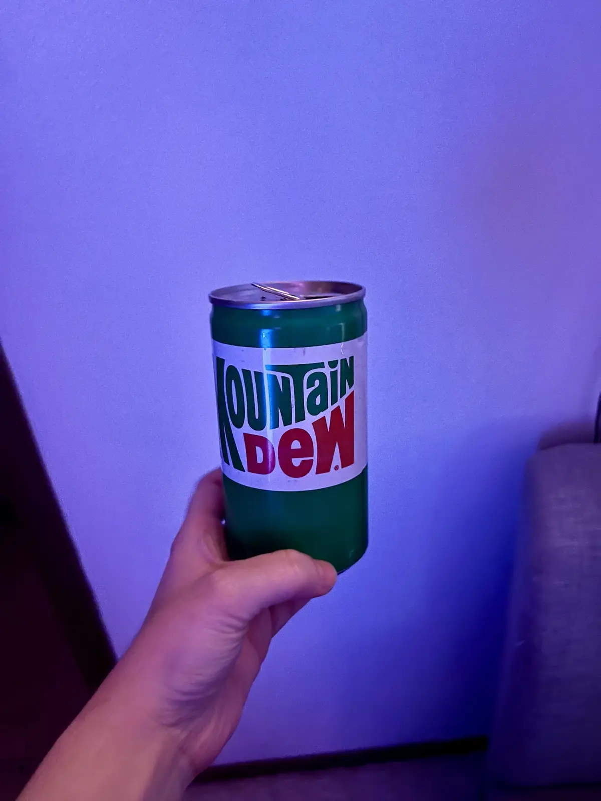 A hand holds an open, green Mountain Dew soda can against a purple-lit wall. The can's label is clearly visible with the…