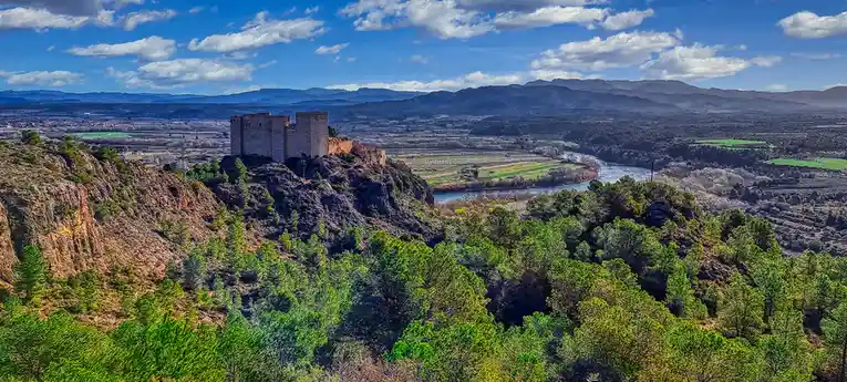 A scenic landscape photo shows a large, ancient stone castle perched atop a rocky hill, overlooking a valley with a winding…