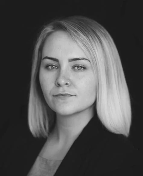 A black-and-white portrait of a woman with blonde shoulder-length hair and a nose ring, looking directly at the camera with…
